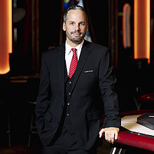 Pokermanager im Casino Graz, Rene Kucher, trägt einen schwarzen Anzug mit rotem Krawatte, steht an einem Pokertisch und lächelt in die Kamera.
